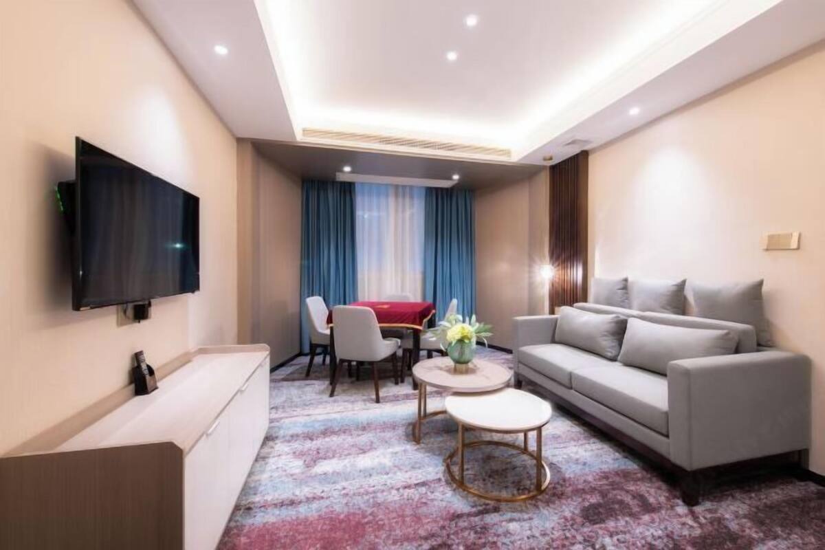 A modern hotel room with a flat-screen TV mounted on the wall, a grey sofa, a small round coffee table, a dining table with chairs, and a patterned carpet.