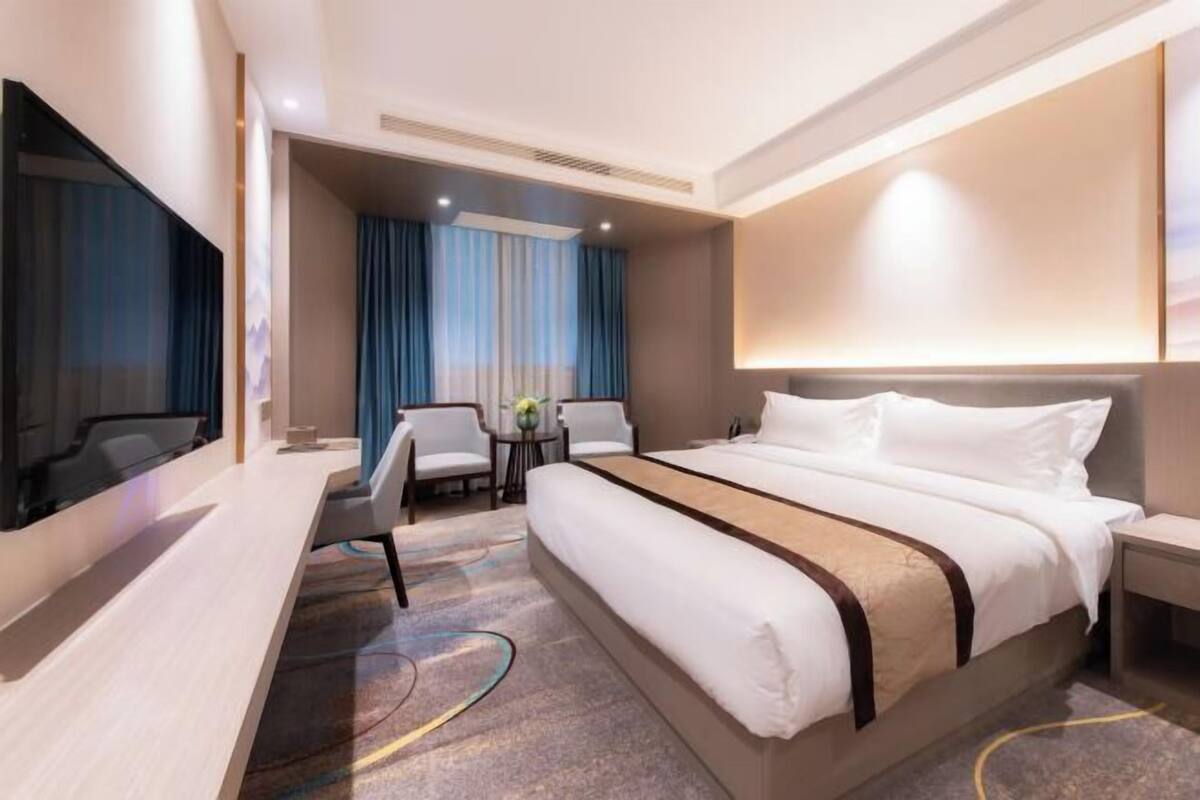 A modern hotel room with a large bed, a desk, a TV, and a seating area with a table and chairs.