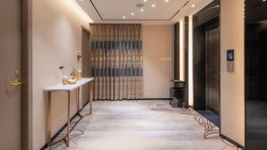 Pasillo de hotel con mostrador de recepción, cortina estampada, ascensor y piso alfombrado.