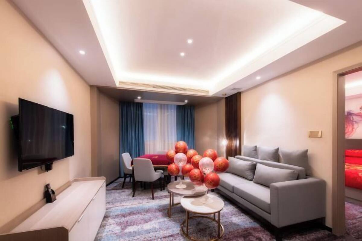 A modern hotel room with a flat-screen TV, a sofa, a dining table with chairs, and a decorative arrangement of red and white spherical objects.