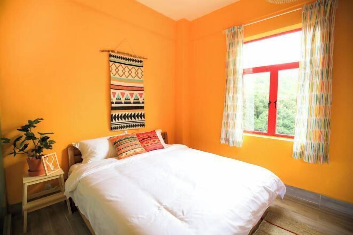 A bedroom with a bed, a window with curtains, a wall hanging, and a small table with a plant.