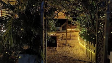 A nighttime view of a garden area with a wooden pergola, string lights, and a small building with a balcony.