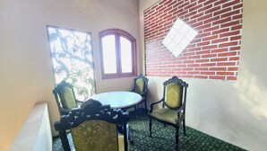 Un petit balcon fermé, avec une table ronde et quatre chaises, une fenêtre agrémentée d’un paravent décoratif et un mur de briques percé d’une fenêtre en forme de losange.