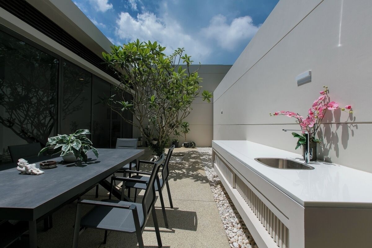 Un espacio moderno para comer al aire libre con una mesa y sillas, rodeado de plantas y un mostrador blanco con un lavamanos.