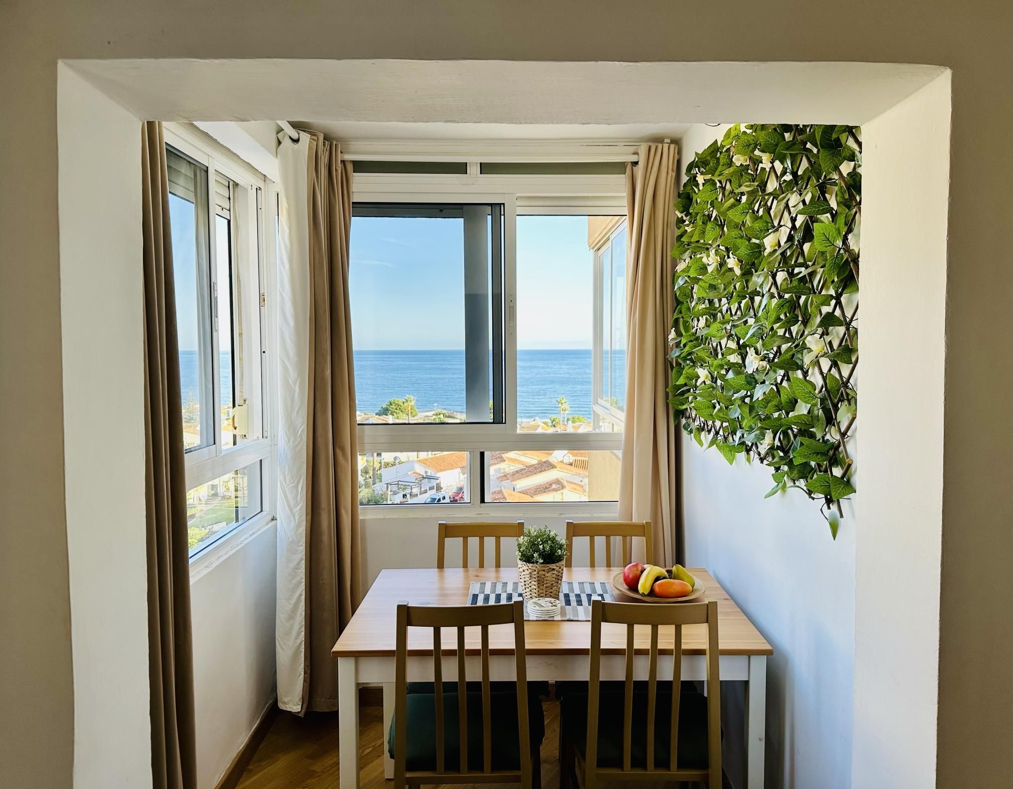 A dining area with a table and chairs, a view of the sea through large windows, and a wall-mounted plant.