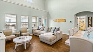 A spacious living room with a high ceiling, large windows, a dining area, and a view of the balcony.