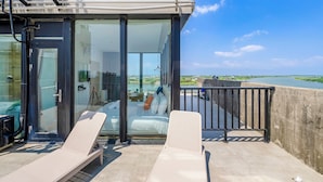 Un balcon avec deux fauteuils, une porte vitrée donnant sur une chambre à coucher avec un lit et des oreillers, et une vue sur une étendue d’eau et la verdure.
