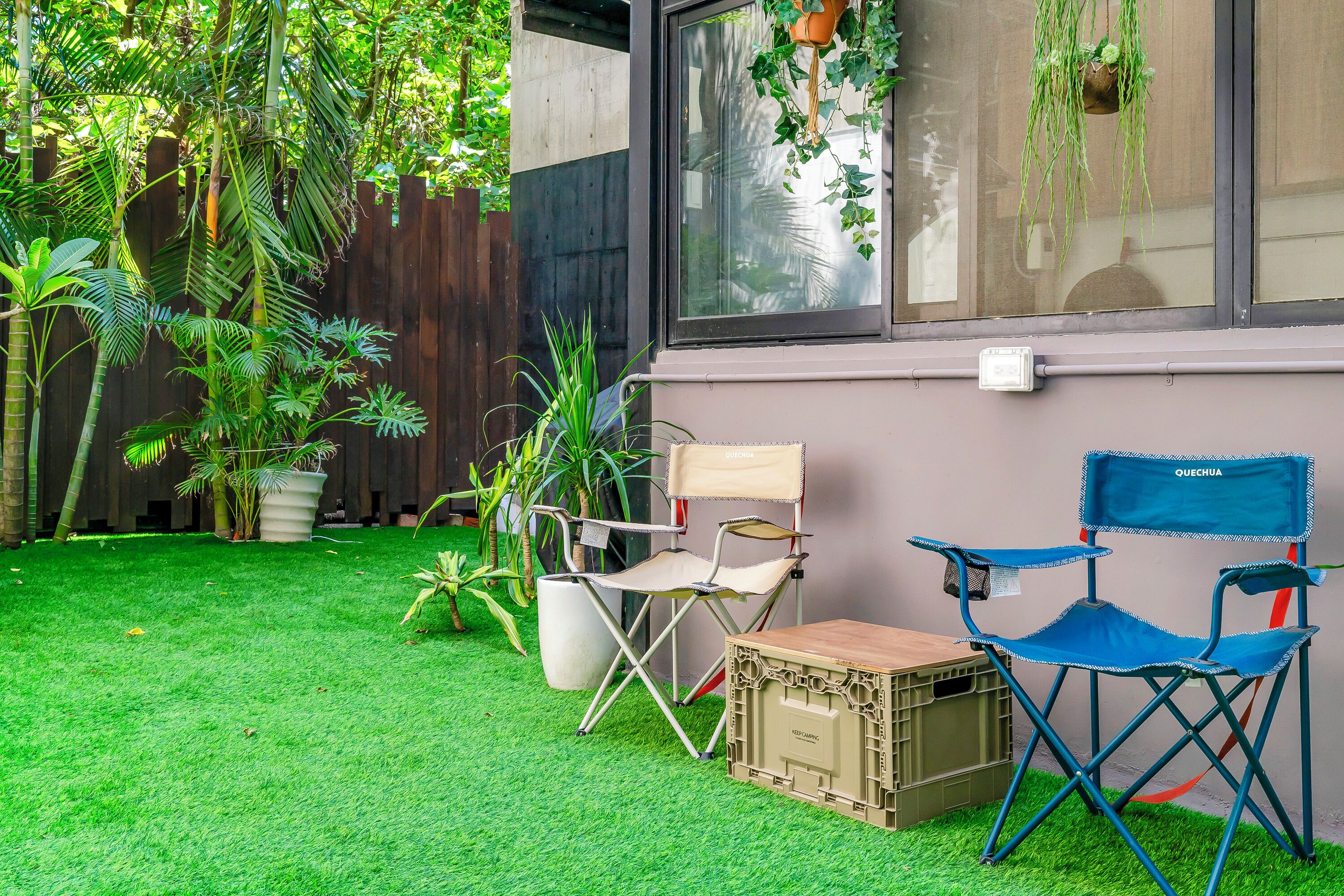 A small outdoor area with artificial grass, two folding chairs, a crate, and a wooden fence.