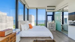 Une chambre d’hôtel moderne avec un grand lit, une commode en bois, une petite table et une vue sur la ville et l’océan.