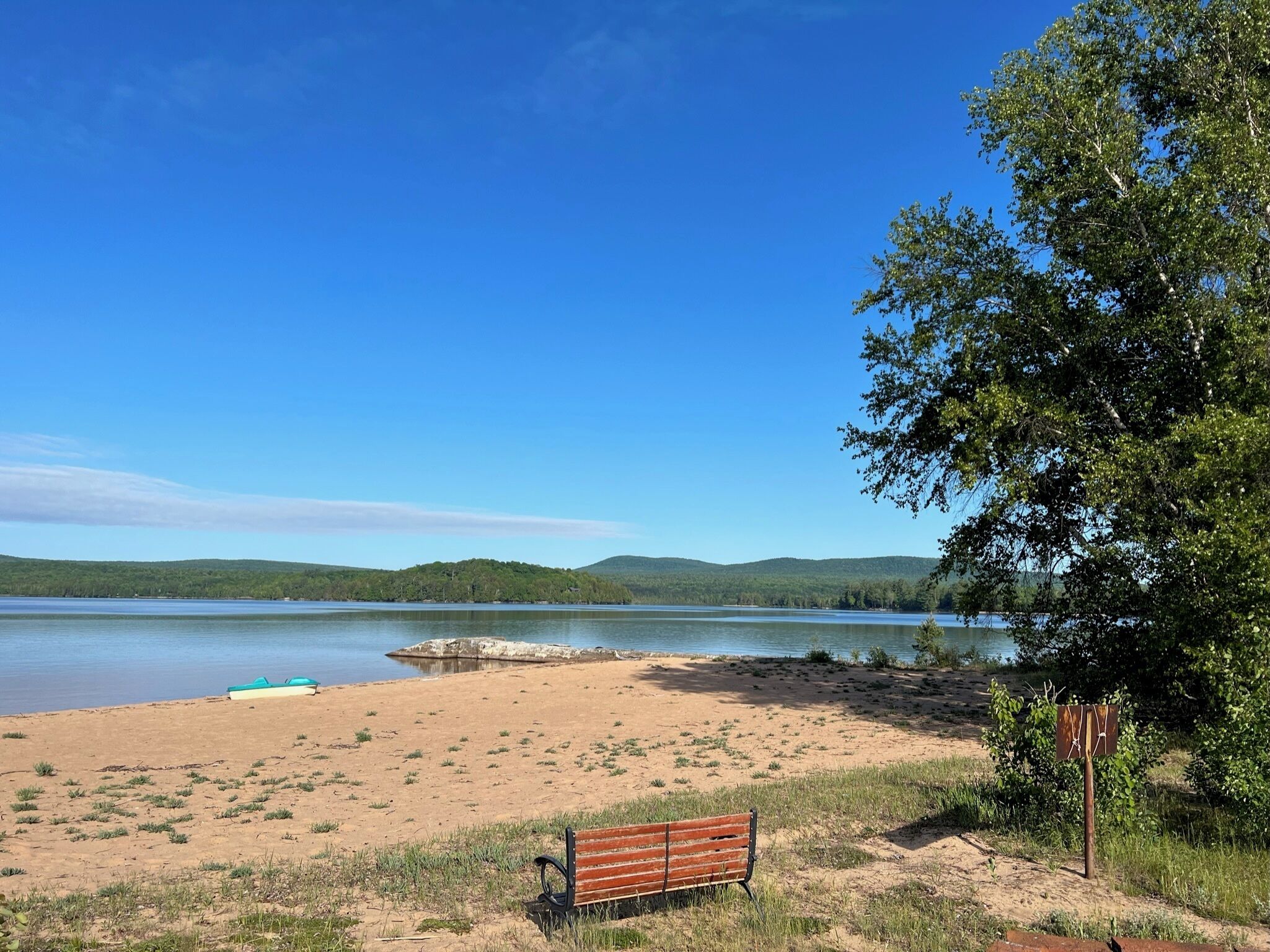 Ein sandiger Strand mit einer Bank und einem Boot, umgeben von Bäumen und Hügeln im Hintergrund.