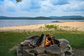 Un feu de camp dans un foyer en pierre, entouré d’une zone herbeuse, avec un lac et des montagnes en arrière-plan.
