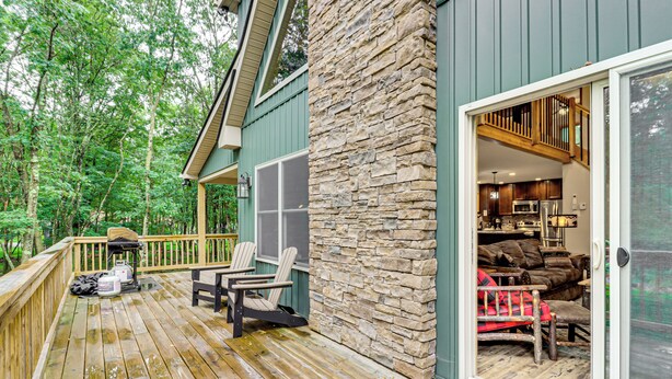 A wooden deck with outdoor seating, a stone wall, and a view of the interior through a glass door. - Forest Views & Hot Tub: Poconos Cozy Cabin (Albrightsville)