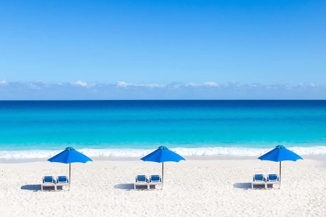 A beach with three blue umbrellas and white lounge chairs.
