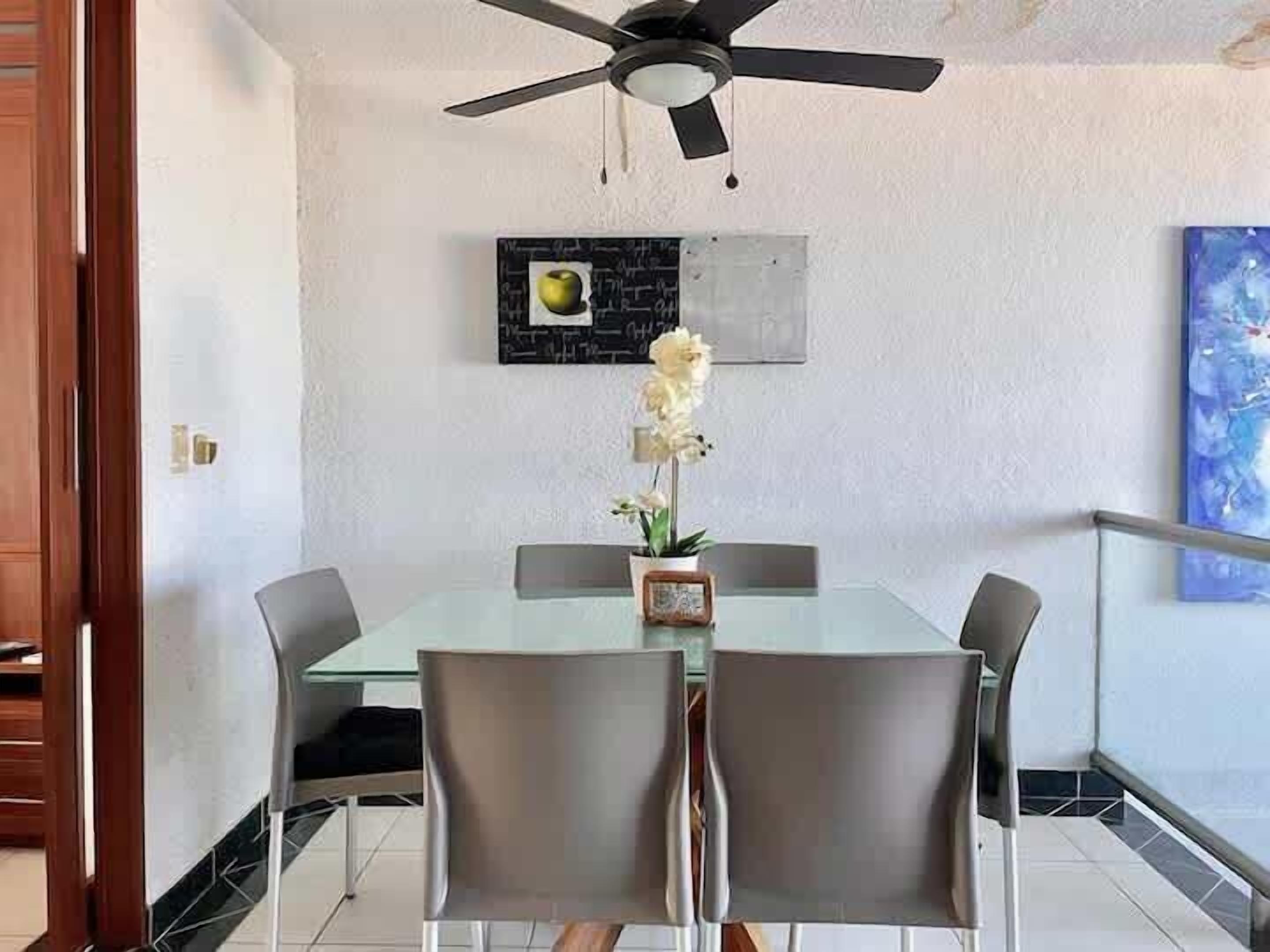A dining area with a glass table, four chairs, a ceiling fan, a framed picture, and a flower vase.
