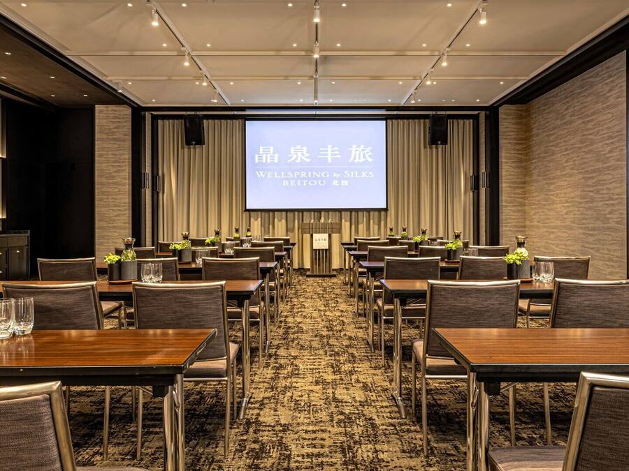 A conference room with a large screen displaying text, multiple tables with chairs, and a carpeted floor.