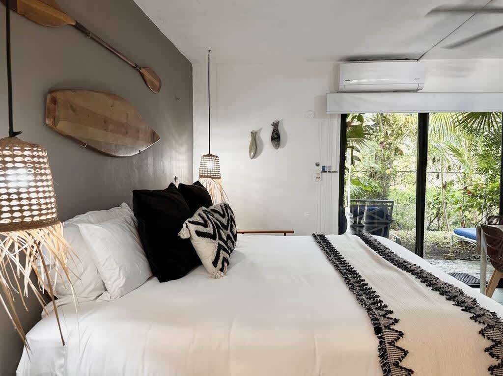 Un dormitorio con cama, almohadas, lámpara colgante, aire acondicionado instalado en la pared y vista a zonas verdes al exterior.
