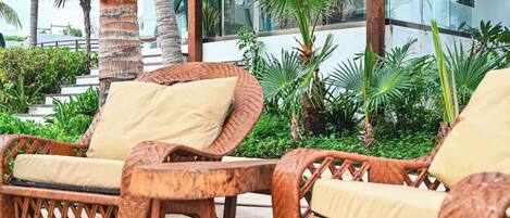 Des fauteuils de jardin en osier, dotés de coussins, dans un décor extérieur agrémenté de palmiers et avec un bâtiment moderne en arrière-plan.