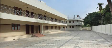 A multi-story building with a flat roof, a balcony on the ground floor, and a paved courtyard.