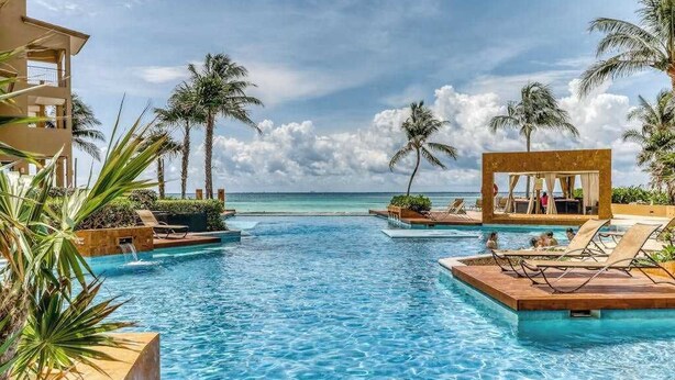 Un espace piscine aménagé avec des chaises longues, des palmiers et une vue sur l’océan.