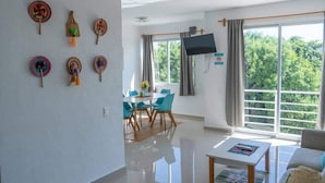 A modern living room with a flat-screen TV, a dining area with blue chairs, and a balcony with a view of trees. - Family Suite | 2BR, 2 Bath, Pool & 24h Security (Cancún)