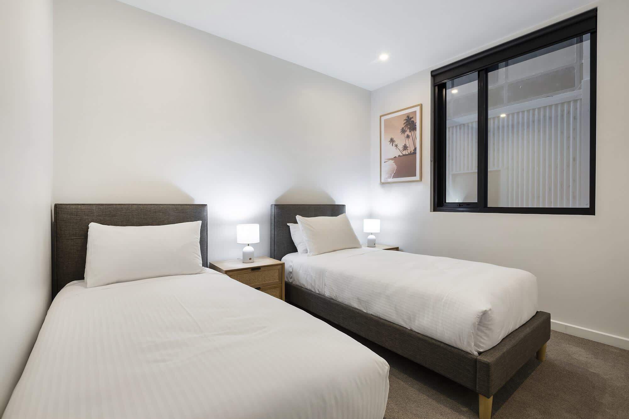 A hotel room with two single beds, bedside tables with lamps, a window with blinds, and a framed picture on the wall.