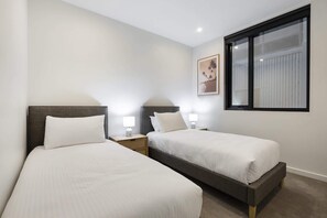 A hotel room with two single beds, bedside tables with lamps, a window with blinds, and a framed picture on the wall. - Oceanfront 2-Bed With Balcony & Study Nook (Geelong)