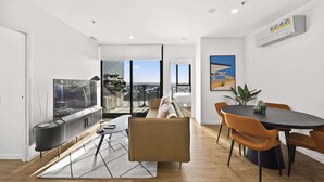 A modern living room with a large window, a sofa, a dining table with chairs, and a radiator. - Refined 2-Bed With Luxurious Building Amenities (Belconnen)