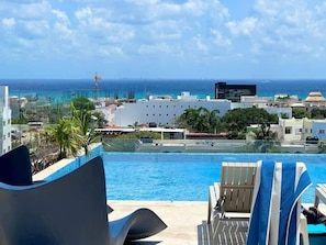 Vista da piscina com espreguiçadeiras, uma visão clara do oceano e edifícios ao fundo.