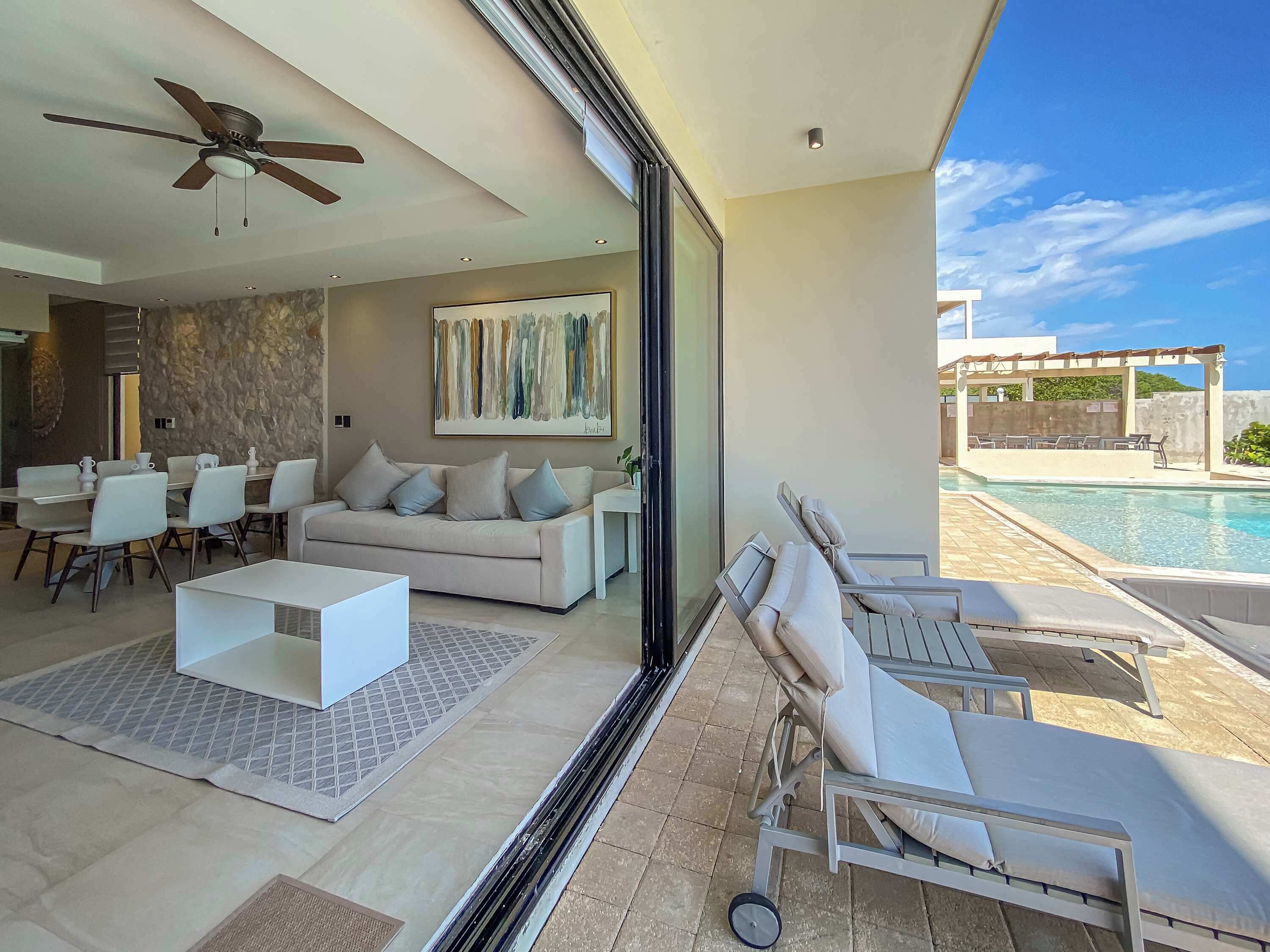 A modern living room with a sofa, coffee table, and chairs, a dining area with a table and chairs, a ceiling fan, and a large window overlooking a pool.