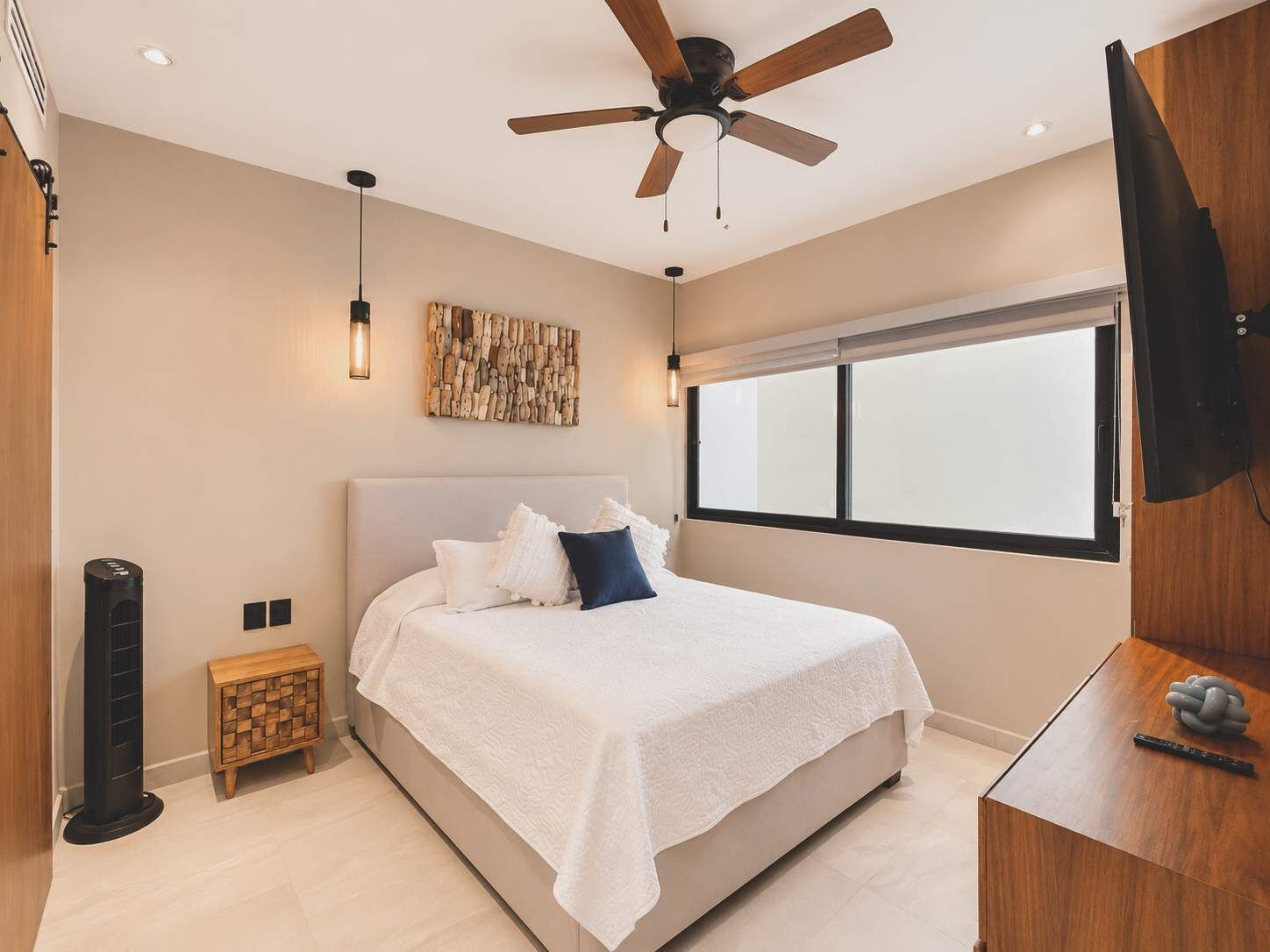 Quarto com cama, ventilador de teto, obra de arte na parede, janela e televisão sobre um suporte de madeira.