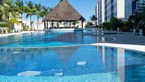 Una piscina con una estructura de techo de paja, palmeras y un edificio moderno al fondo.