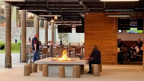 Un área de estar al aire libre con una chimenea central, taburetes de madera y un suelo de baldosas de piedra.