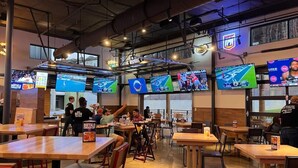 Un restaurante con varias pantallas mostrando eventos deportivos, mesas y sillas de madera, y un ambiente informal para comer.