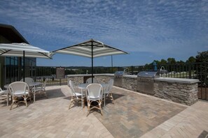 Eine Terrrasse mit einem Grillplatz aus Stein, weißen Korbmöbeln und einem großen Sonnenschirm.