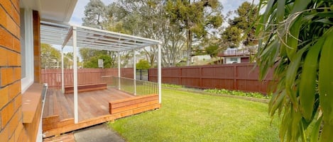 A covered outdoor area with a wooden deck, a fenced yard, and a grassy lawn.