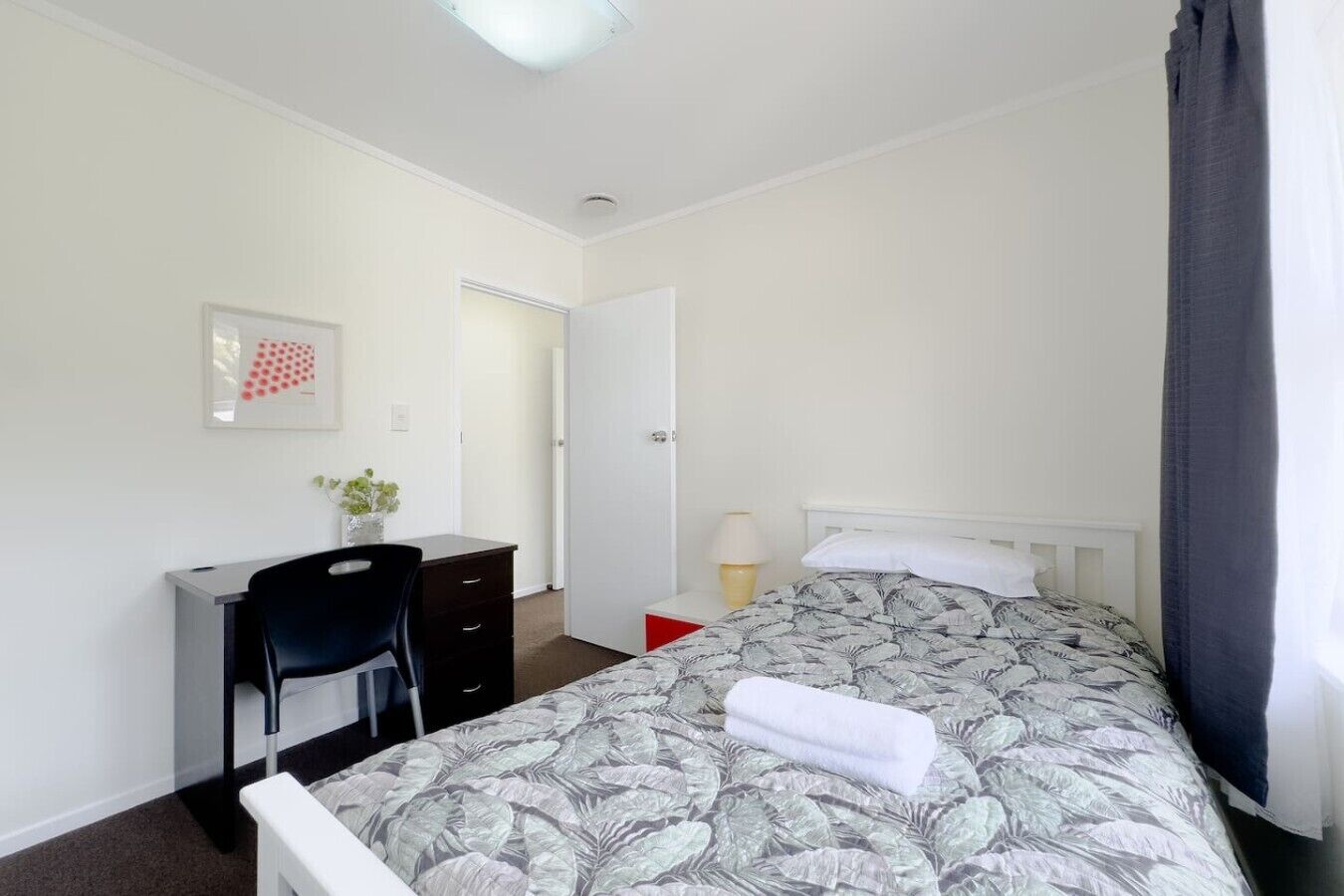 A bedroom with a bed, a desk, a chair, a door, and a small table with a lamp.