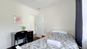 A bedroom with a bed, a desk, a chair, a door, and a small table with a lamp.
