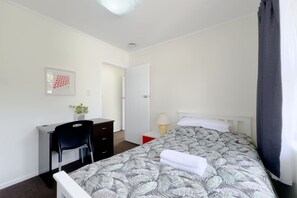 A bedroom with a bed, a desk, a chair, a door, and a small table with a lamp.