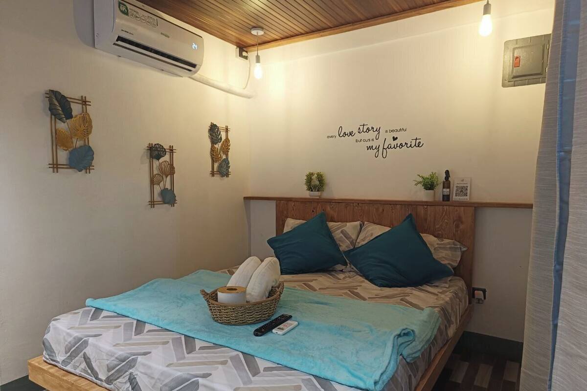 Cama bem arrumada com manta azul, cesta de artigos de higiene pessoal e parede com uma frase decorativa.
