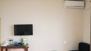A room with a flat-screen TV mounted on the wall, a small round table with a green coffee maker, a blue chair, and an air conditioning unit. - Indrasthan himalaya Resort  (Chandragiri)