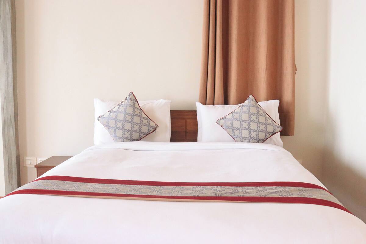 A hotel room with a neatly made bed, two pillows with a geometric pattern, a red and gray bedspread, and a brown curtain.