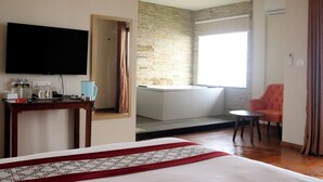 A hotel room with a bed, a TV, a small table with a lamp, a chair, a bathroom with a bathtub, and a large window. - Indrasthan himalaya Resort  (Chandragiri)