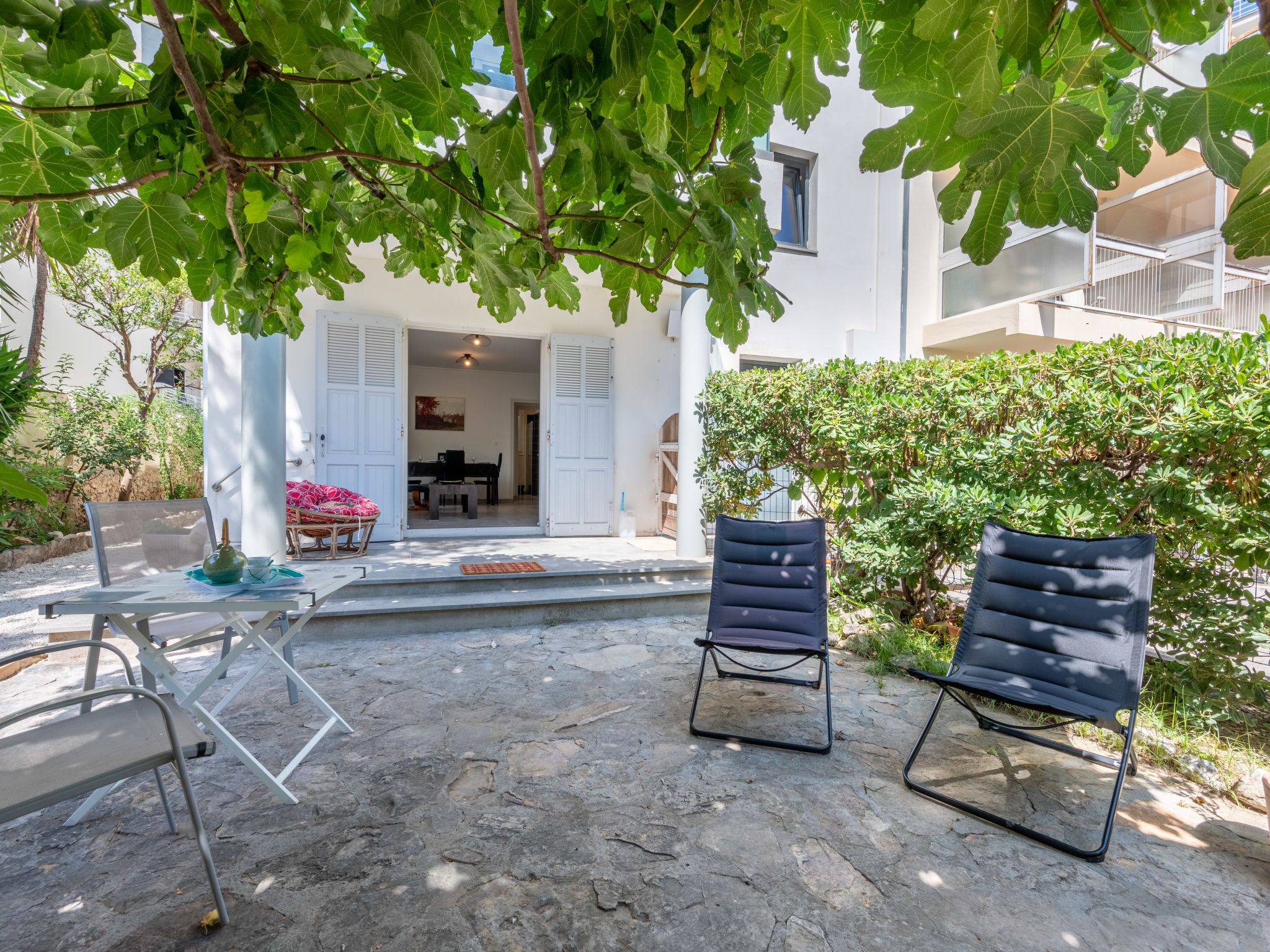 A courtyard with stone flooring, two black lounge chairs, a dining area with a table and chairs, and a white building with shutters.