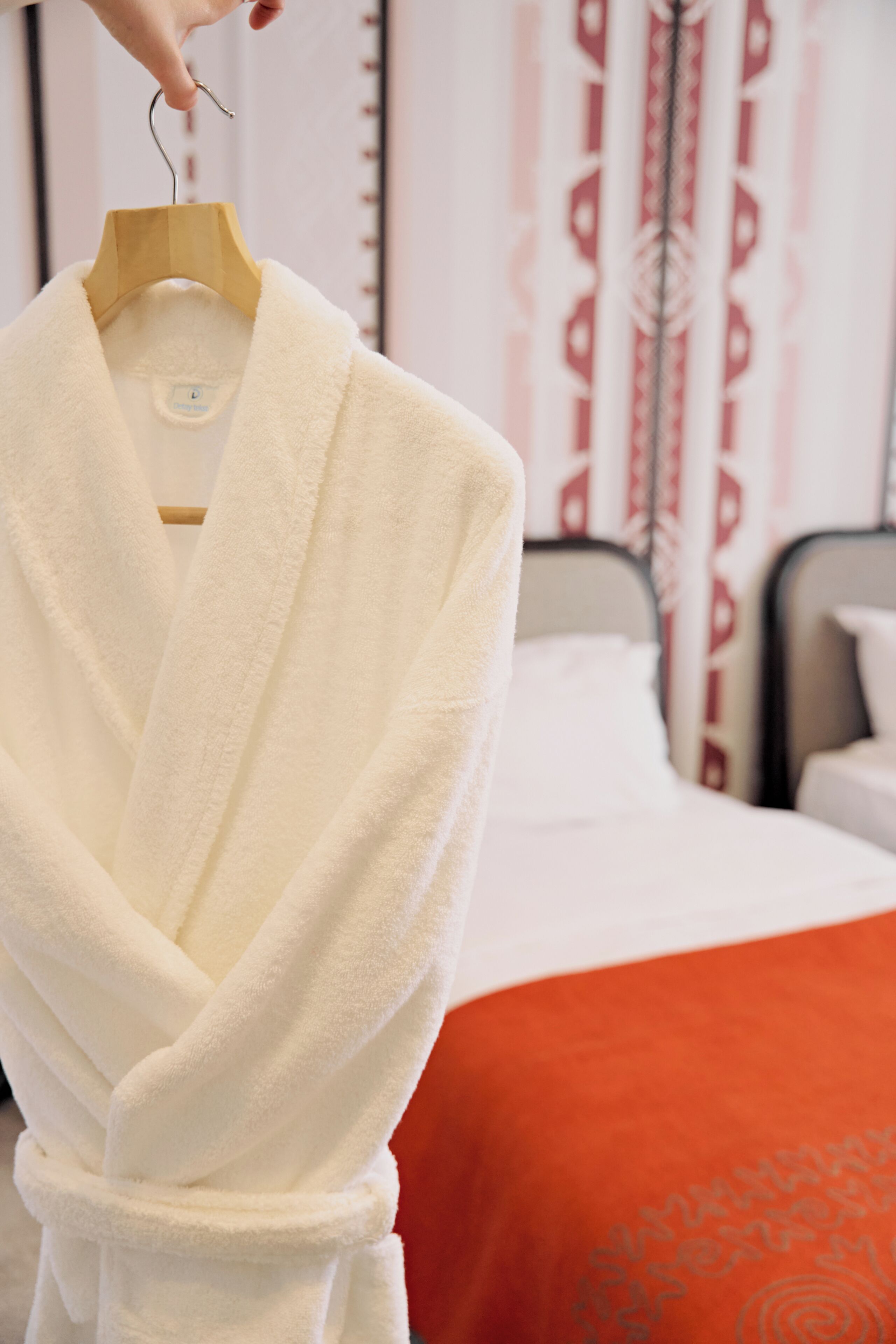 A white bathrobe on a wooden hanger in a hotel room with a bed and a red blanket.