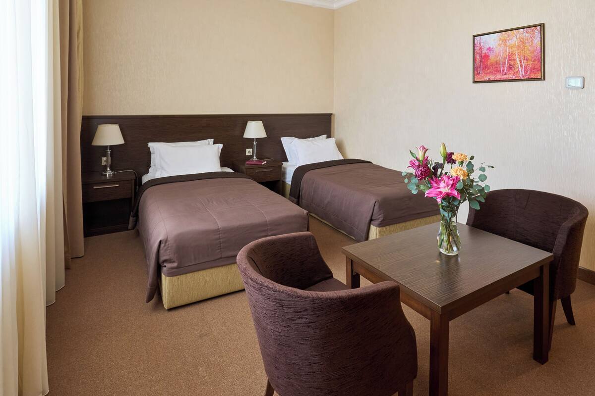 A hotel room with two beds, a table, and chairs. There is a vase with flowers on the table.