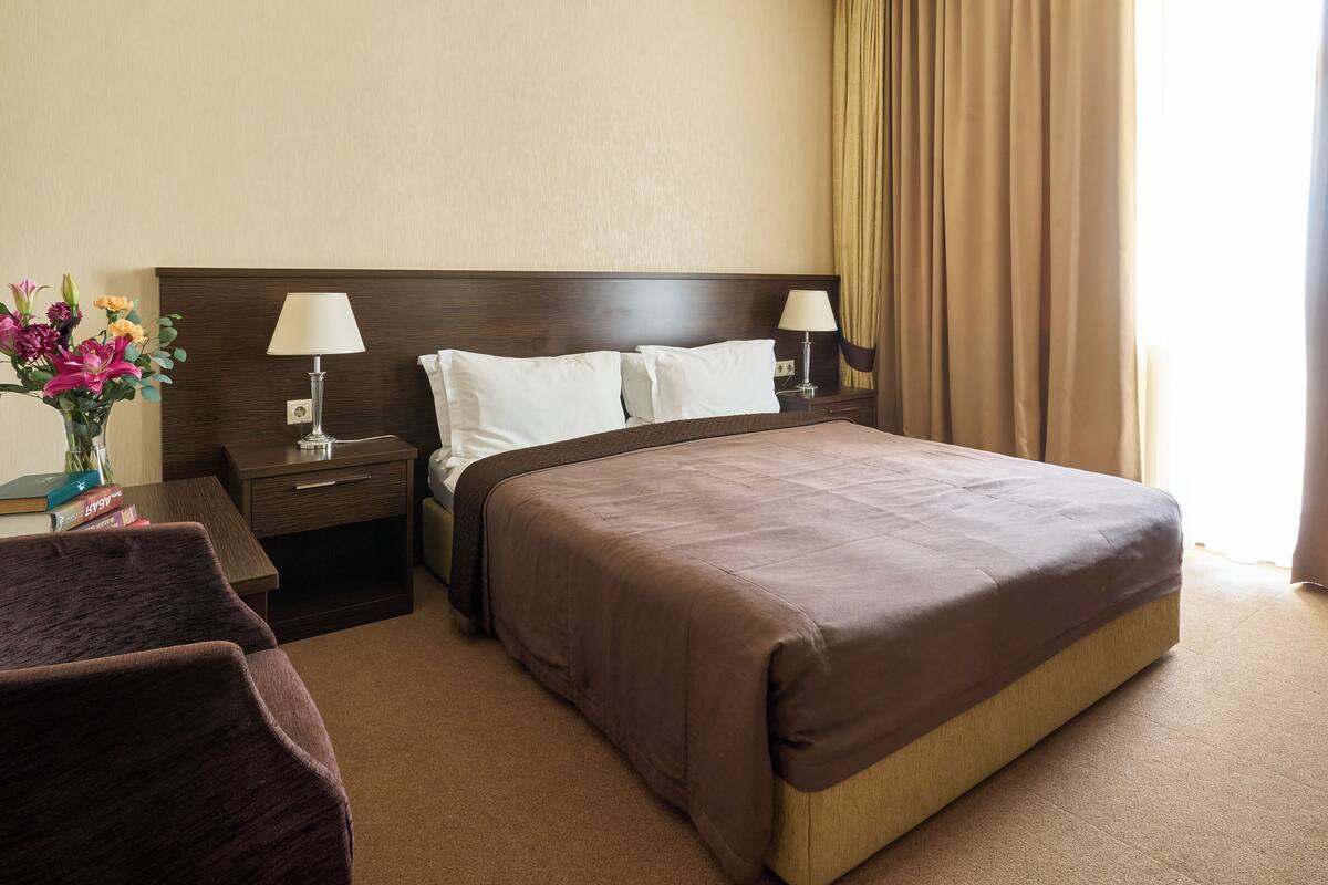 A hotel room with a large bed, two bedside tables with lamps, a nightstand, a chair, and a vase with flowers.