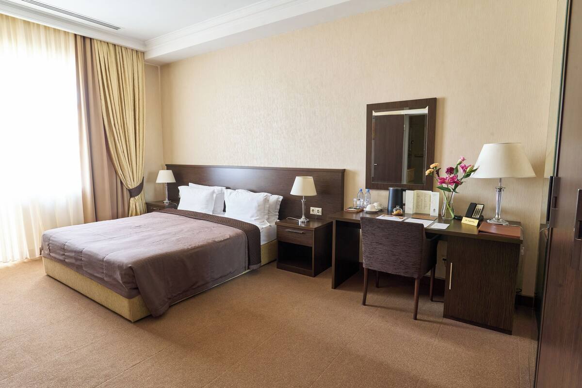 A hotel room with a large bed, a desk with a chair, a nightstand, a mirror, and a lamp.