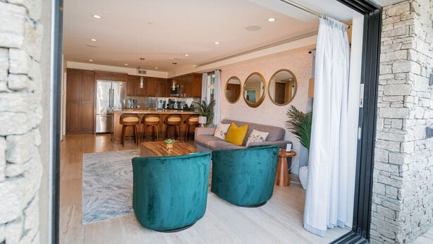 A modern living room with teal armchairs, a wooden coffee table, and a sofa with cushions. A kitchen with wooden cabinets and a dining area with bar stools is visible in the background. - 3 BR, Roof Deck, Ocean Views, Hot Tub, Beach Block (Newport Beach)