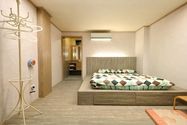A bedroom with a bed, a wooden floor, a wall-mounted air conditioner, and a hallway leading to a bathroom.