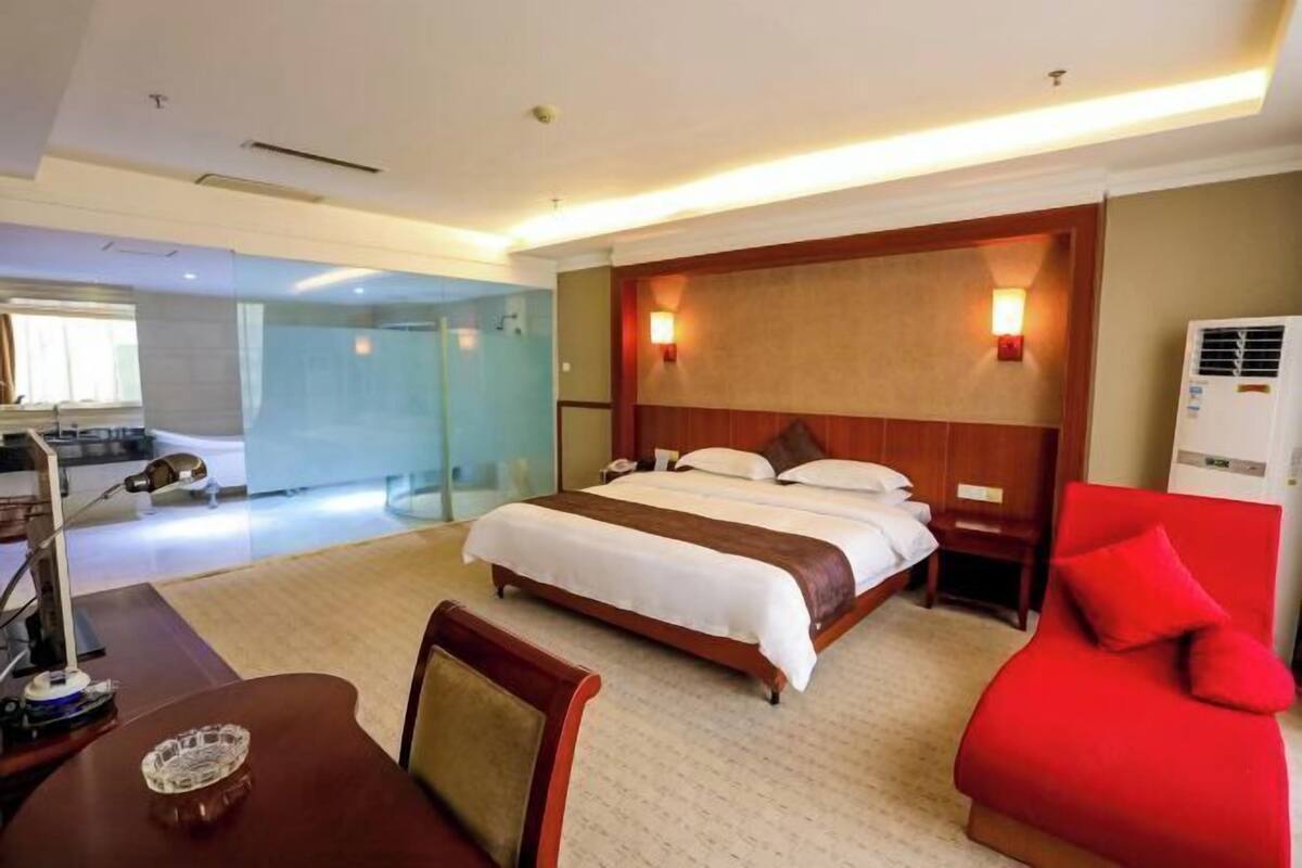A hotel room with a bed, a desk, a chair, a red sofa, and a glass partition.